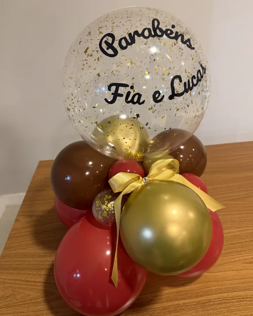 Arranjo de balões vermelhos, marrom chocolate e dourados com balão bubble de confete dourado e mensagem “Parabéns”